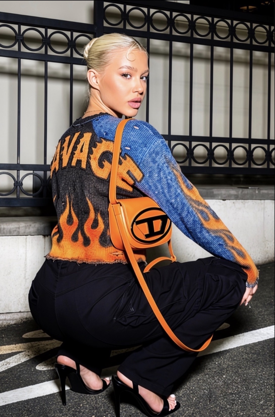 Model crouching by a black metal fence wearing the orange and blue SAVAGE flame knit